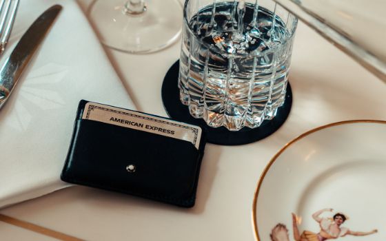 Eine silberne Kreditkarte von American Express aus Metall steckt in einem schwarzen Lederetui neben einem Wasserglas auf einem elegant gedeckten Tisch.