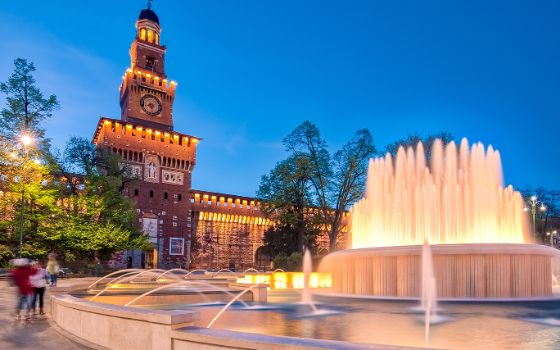 Beleuchtetes Castello Sforzesco in Mailand bei Abenddämmerung mit Springbrunnen im Vordergrund und blauem Himmel im Hintergrund.