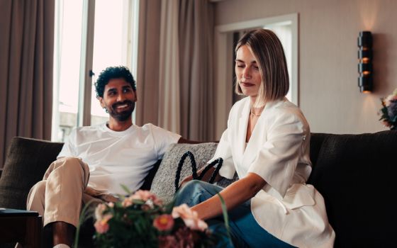 Ein Mann und eine Frau sitzen auf einem Sofa in einem Hotelzimmer, er schaut lächelnd auf sie, während sie sich zu dem Tisch vor dem Sofa beugt. 