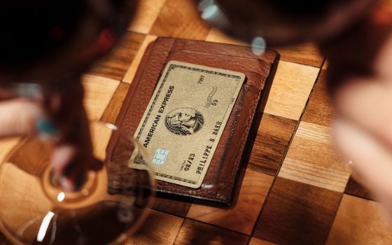 Eine goldene Kreditkarte von American Express auf einem braunen Lederetui aus auf einem gemusterten Holztisch, gerahmt von zwei gefüllten Rotweingläsern