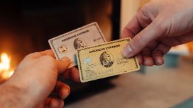Eine goldene Kreditkarte von American Express wird von einer Hand vor eine silberne Kreditkarte von American Express in einer anderen Hand gehalten, im Hintergrund ein Kaminfeuer in Unschärfe.