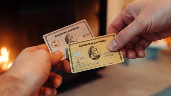 Eine goldene Kreditkarte von American Express wird von einer Hand vor eine silberne Kreditkarte von American Express in einer anderen Hand gehalten, im Hintergrund ein Kaminfeuer in Unschärfe.