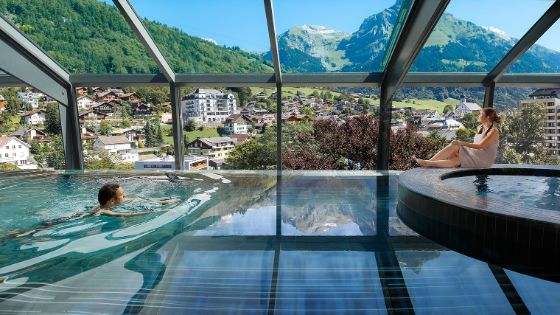 Ein Mann schwimmt in einem Pool und eine Frau sitzt am Rand unter einem Glasdach mit Blick in die Berge.