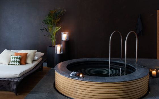 Eine gepolsterte Liege vor einer dunklen Wand. Daneben brennende Kerzen und ein holzverkleidetes Becken.