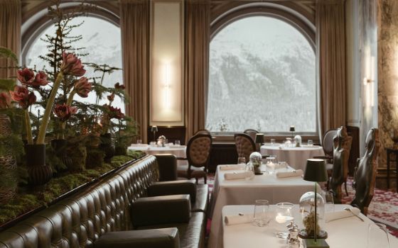 Eleganter Speiseraum mit Teppichboden und Ledersesseln an weiss eingedeckten Tischen mit Blick auf schneebedeckte Berglandschaft durch grosse Bogenfenster.