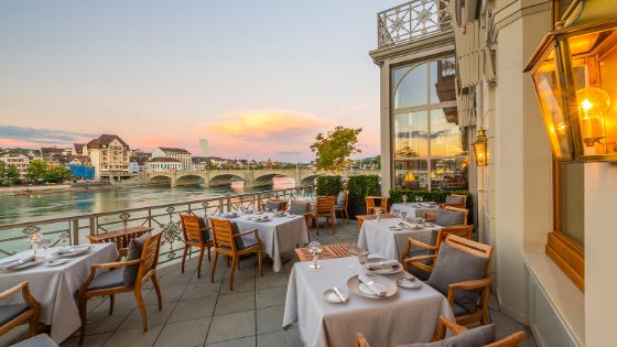 Weiss eingedeckte Tische auf einer Terrasse eines luxuriösen Gebäudes am Fluss mit Blick auf eine Bogenbrücke und Basler Stadtpanorama. 