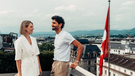 Ein Paar steht lächelnd auf einer Dachterrasse neben einer Schweizer Flagge, im Hintergrund Dächer.