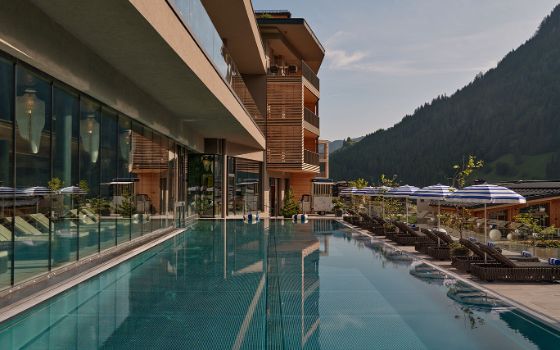 Hotelterrasse mit Pool und Sonnenliegen vor einem bewaldeten Berg.
