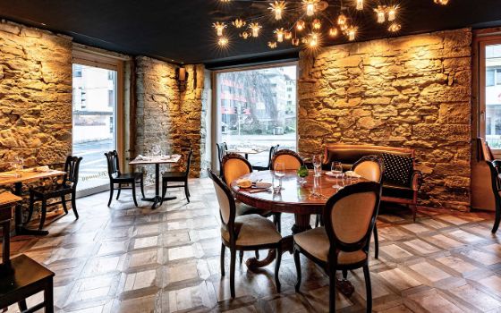 Eingedeckte Holztische in einem eleganten Speiseraum mit Parkettboden, angeleuchteten Steinmauern und bodentiefen Fenstern in urbaner Umgebung.