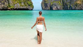 Rückansicht einer Frau in Strandkleidung, die an einem weissen Sandstrand durchs flache Wasser läuft vor bewachsenen Kalksteinfelsen im türkisblauen Meer. 