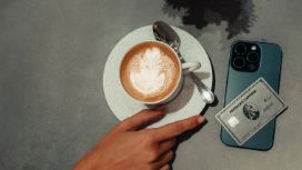 Eine Hand greift nach einer Tasse Cappuccino, daneben liegt eine Kreditkarte von American Express auf einem Smartphone.