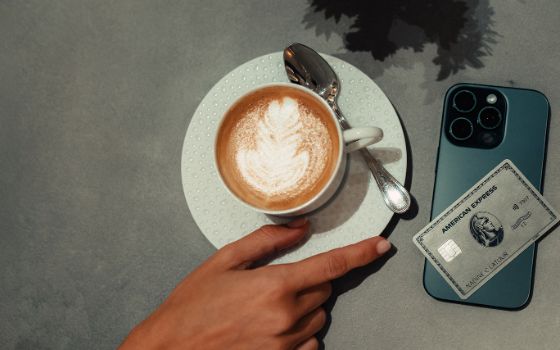 Eine Hand greift nach einer Tasse Cappuccino, daneben liegt eine Kreditkarte von American Express auf einem Smartphone.