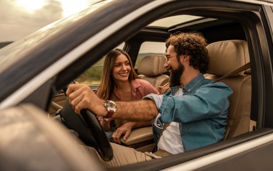 Eine Frau und ein Mann sitzen in einem Auto und fahren eine Strasse entlang, er fährt und schaut lächelnd zu ihr. 