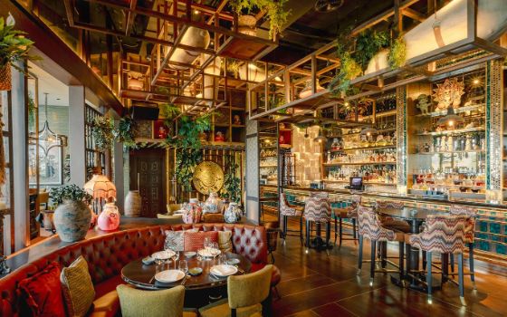 In einem Restaurant stehen ein rotes, ovales Ledersofa und Stühle um einen Tisch, an der Wand befindet sich eine Bar mit grosser, verzierter Vitrine und von der Decke hängen Pflanzen und hölzerne Gestelle. 