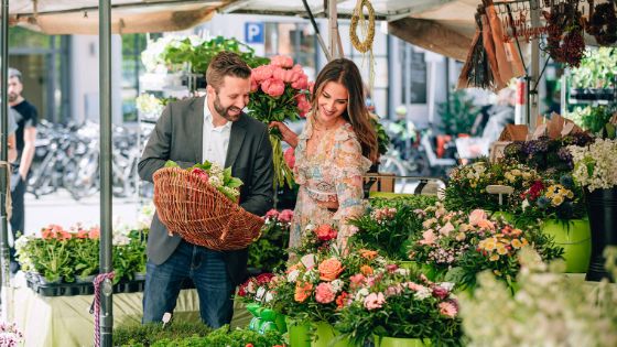 Ein lächelndes Paar mit Einkaufskorb an einem Marktstand mit Blumen in Innenstadtlage. 
