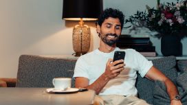 Ein Mann im weissen T-Shirt sitzt entspannt auf einem Sofa an einem Tisch mit Kaffeetasse und schaut lächelnd auf ein Smartphone in seiner Hand.