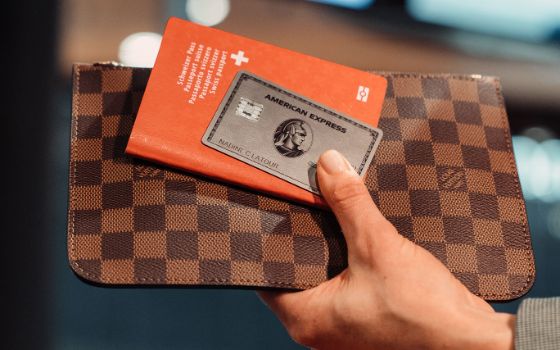 Eine Hand hält eine silberne Kreditkarte von American Express auf einem roten Reisepass auf einem braunen Lederetui hoch. 
