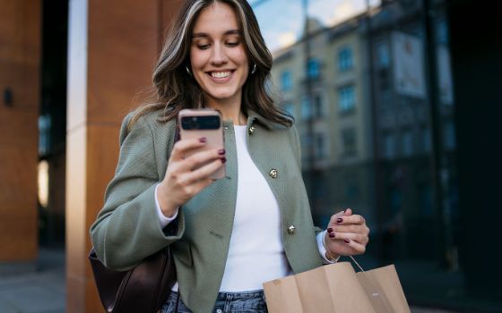 Eine junge, elegant gekleidete Frau steht mit einer Einkaufstüte in der Hand in urbaner Umgebung und schaut lächelnd auf ihr Smartphone.