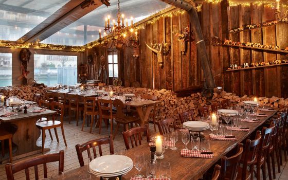 Lange Holztische und Stühle stehen in einem Raum, die Wände sind ebenfalls aus Holz und an den Wänden hängt Holz-Dekoration.