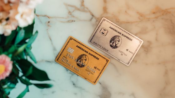 Zwei American Express Karten, eine goldene und eine platinfarbene, liegen auf einer hellen Marmorfläche. Links befindet sich ein unscharfer Blumenstrauss im Vordergrund.