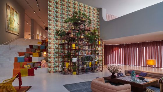 Mittig in einem Raum steht eine bunte Wand vor der ein grosses Regal mit unterschiedlichen Pflanzen und Vasen platziert sind. Auf der linken Seite geht eine hellgraue Treppe zur nächsten Etage, auf der verschiedene, bunte Teppiche und Kissen liegen.