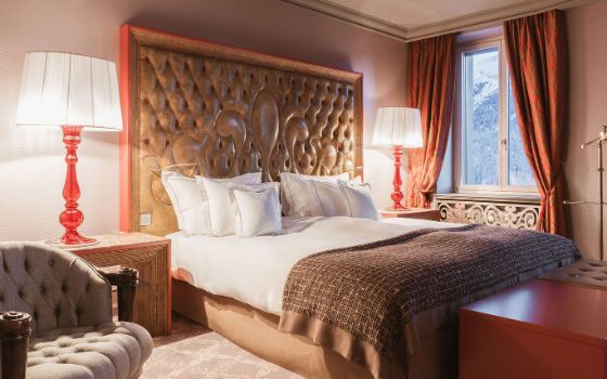 Elegantes Schlafzimmer mit Doppelbett mit einem hohen, verzierten Kopfteil aus Leder, übergrossen Nachttischlampen und Blick in Berglandschaft.