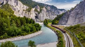 Ein roter Zug fährt auf Gleisen entlang eines Flusses mit hellblauem Wasser in einem bewaldeten Tal zwischen steilen Felswänden. 
