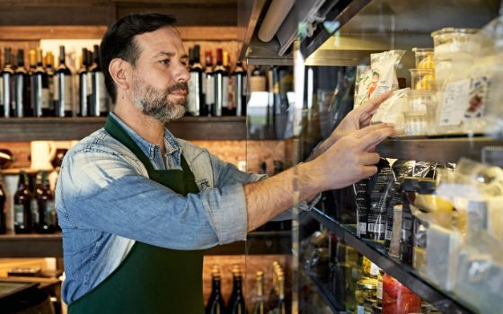 Ein Mann in Jeanshemd und dunkler Schürze hantiert mit Lebensmitteln in einer Vitrine, im Hintegrund Weinflaschen und dunklen Holzregalen.