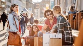 Mehrere gut gelaunte Personen sitzen mit Einkaufstaschen auf einer Treppe in einer belebten Fussgängerzone in einer Altstadt bei Sonnenschein. 
