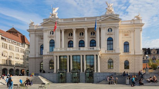 Aussenfassade des historischen Opernhauses in Zürich, davor sitzen Personen auf Treppenstufen.