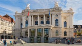 Aussenfassade des historischen Opernhauses in Zürich, davor sitzen Personen auf Treppenstufen. 