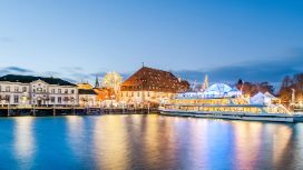 Erleuchteter Weihnachtsmarkt mit Schiff am Ufer eines Sees unter blauem Himmel am Abend. 