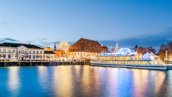 Erleuchteter Weihnachtsmarkt mit Schiff am Ufer eines Sees unter blauem Himmel am Abend. 