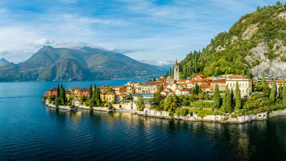 Mediterraner Ort mit Zypressen am Fuße eines Berges an einem See, im Hintergrund Bergpanorama.