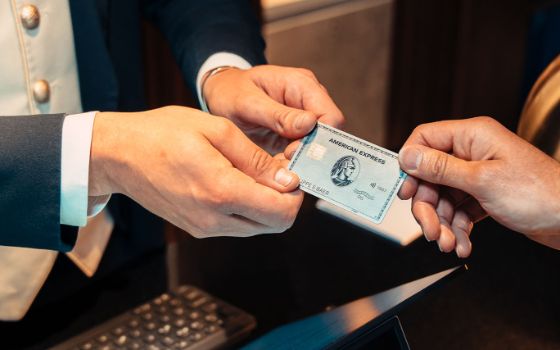 Eine silberne Kreditkarte von American Express wird von einer Hand in die Hände einer elegant gekleideten Person an einer Rezeption übergeben.