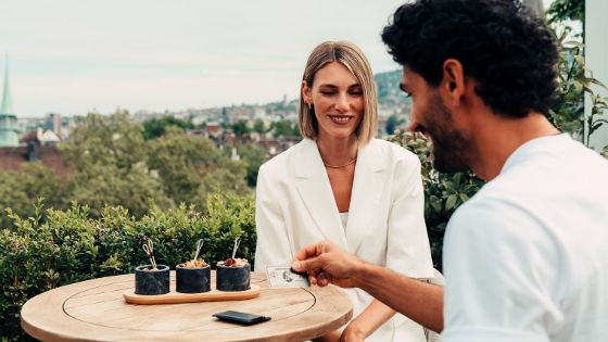 Ein Paar sitzt an einem Tisch auf einer Terrasse mit Stadtblick, er hält eine silberne Kreditkarte von American Express in der Hand.