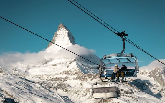 Zwei Skifahrende sitzen in einem Sessellift vor schneebedecktem Bergpanorama mit spitzer Felsformation unter blauem Himmel.