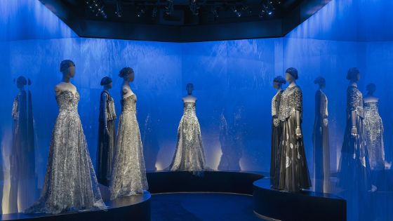 In einem Raum mit blauen Wänden stehen sechs Schaufensterpuppen mit hellen, glitzernden Kleidern. 