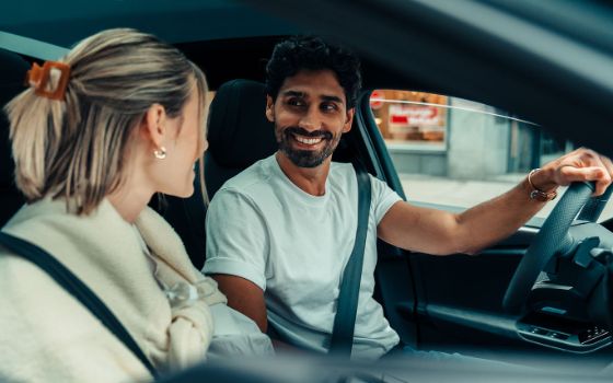 Ein angeschnalltes Paar auf den Vordersitzen eines Autos in urbaner Umgebung schaut sich lächelnd an, er hat dabei die Hand am Steuer.
