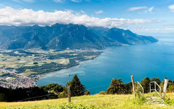 Blick auf den Genfersee mit den Alpen im Hintergrund, aufgenommen von einem Aussichtspunkt oberhalb der Lavaux-Region bei sonnigem Wetter.