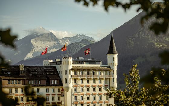 Luxuriöser Hotelkomplex in schlossartigem Architekturstil mit Rundturm und wehenden Fahnen vor Bergpanorama, im Vordergrund grüne Baumzweige. 