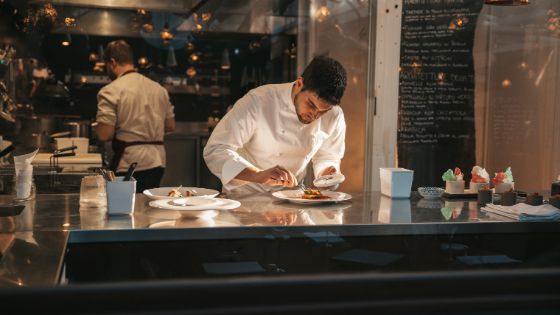 Zwei Köche in einer Restaurantküche, von denen einer weisse Teller, die auf einer Arbeitsplatte stehen, vorsichtig anrichtet.