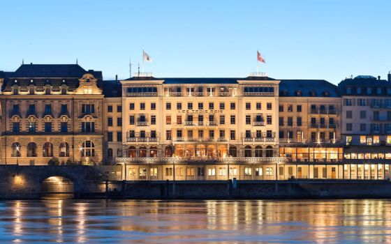 Frontalansicht eines beleuchteten Grand Hotels mit Schweizer Fahnen am Fluss bei Abenddämmerung.