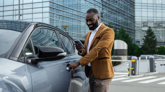 Ein elegant gekleideter Mann schaut lächelnd auf sein Handy und öffnet mit der anderen Hand die Tür eines glänzenden Autos. Im Hintergrund ein verglastes Gebäude.