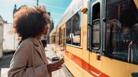 Eine Frau steht am Bahnsteig, als ein Zug einfährt. In der Hand hält sie einen Coffee-to-go-Becher und eine Kreditkarte von American Express.
