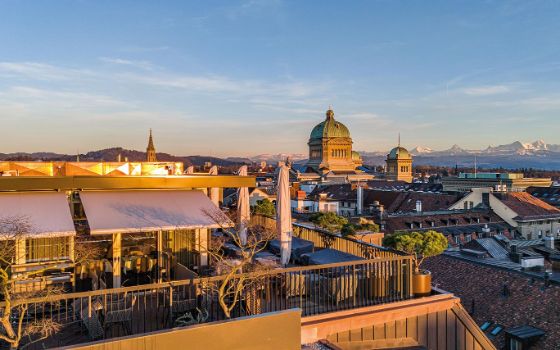 Eine grosse Dachterrasse mit Möbeln und Pflanzen, im Hintergrund sieht man die Dächer einer Stadt und Berge. 