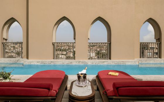 Zwei rote, luxuriöse Sonnenliegen direkt am Rand eines Pools auf einer Dachterrasse. Zwischen den Liegen steht eine Champagnerflasche im Kühler auf einem Beistelltisch, im Hintergrund der Blick durch orientalische Mauerbögen auf ein Stadtpanorama. 