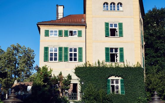Ein gelbes Herrenhaus mit grünen Fensterläden, umgeben von Hecken und Bäumen unter blauen Himmel.