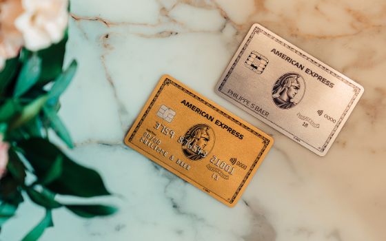 Eine goldene und eine silberne Kreditkarte von American Express auf einem hellen Marmortisch neben einem Blumenstrauss im Anschnitt