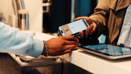 Eine Person hält an einem Tresen ein Smartphone, auf dessen Bildschirm eine American Express Kreditkarte abgebildet ist, an ein Kartenlesegerät, das ihr eine andere Person hinhält. 